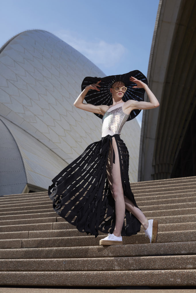 Woman standing on the steps of the Sydney Opera House wearing sunglasses and a large brimmed hat which she holds off her face with both hands.